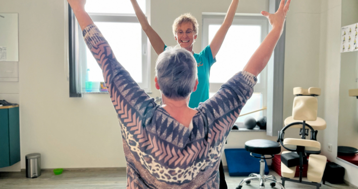 Physiotherapeutin macht gemeinsam mit einer Patientin eine Übung mit erhobenen Armen im Therapieraum.