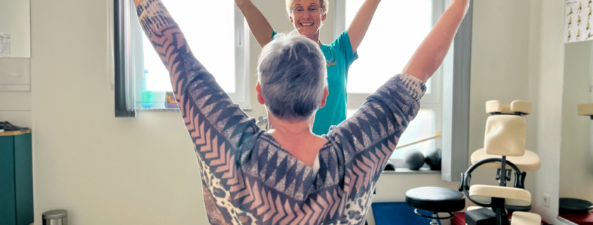 Physiotherapeutin macht gemeinsam mit einer Patientin eine Übung mit erhobenen Armen im Therapieraum.