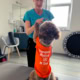 Frau in einem Therapieraum gibt einem kleinen Hund im orangen Shirt ein Kommando oder Leckerli.“