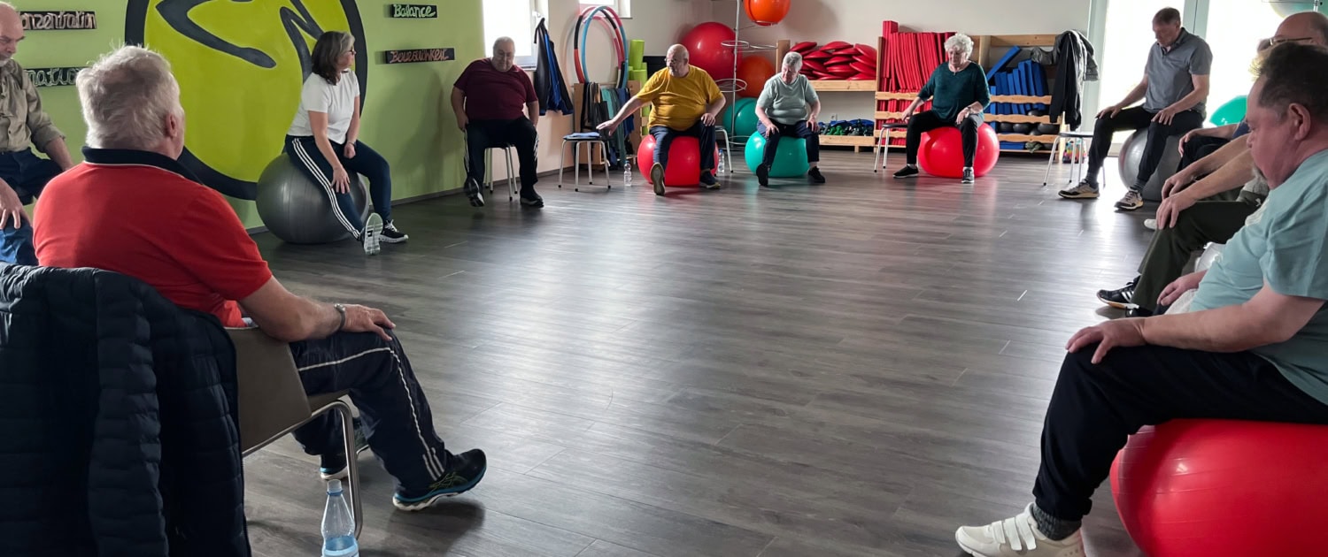 Gruppe älterer Menschen sitzt im Kreis auf Gymnastikbällen und Stühlen während einer Rehasport-Übungseinheit in einem Trainingsraum.